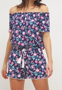 Jumpsuit floreale blu scuro off-the-shoulder, caratterizzato da fiori rosa e turchesi, banda elastica e dettagli con frange in vita e all'orlo.