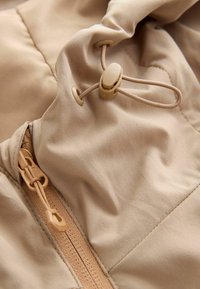 Beige stof met een rits en verstelbaar trekkoortje met koordstopper aan een jas of tas voor het strakker aantrekken en een veilige sluiting.