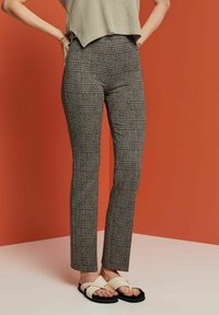 Grå rutiga byxor med en figurnära passform, hög midja och textilstruktur. Bärs med svarta sandalskor. Bakgrunden är en enhetlig orange färg.