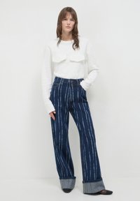 Maglia bianca a maniche lunghe con texture e due tasche sul petto, abbinata a pantaloni in denim blu scuro a vita alta con strisce verticali azzurre.