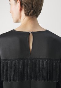 Vestito nero con un profondo taglio sulla schiena, ornato da lunghe frange e dettagli in perline decorative, realizzato in un tessuto leggermente lucido per aggiungere texture.