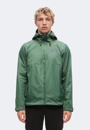 Joven con cabello rubio corto y rizado que lleva una chaqueta impermeable verde con capucha de Patagonia y pantalones negros de pie frente a un fondo liso.