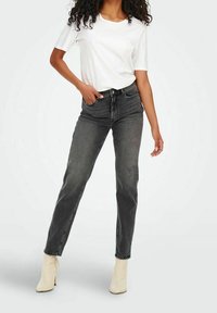 Jean noir taille haute avec une coupe droite, présentant une texture délavée, un design à cinq poches, associé à un t-shirt blanc uni et des bottes beige.