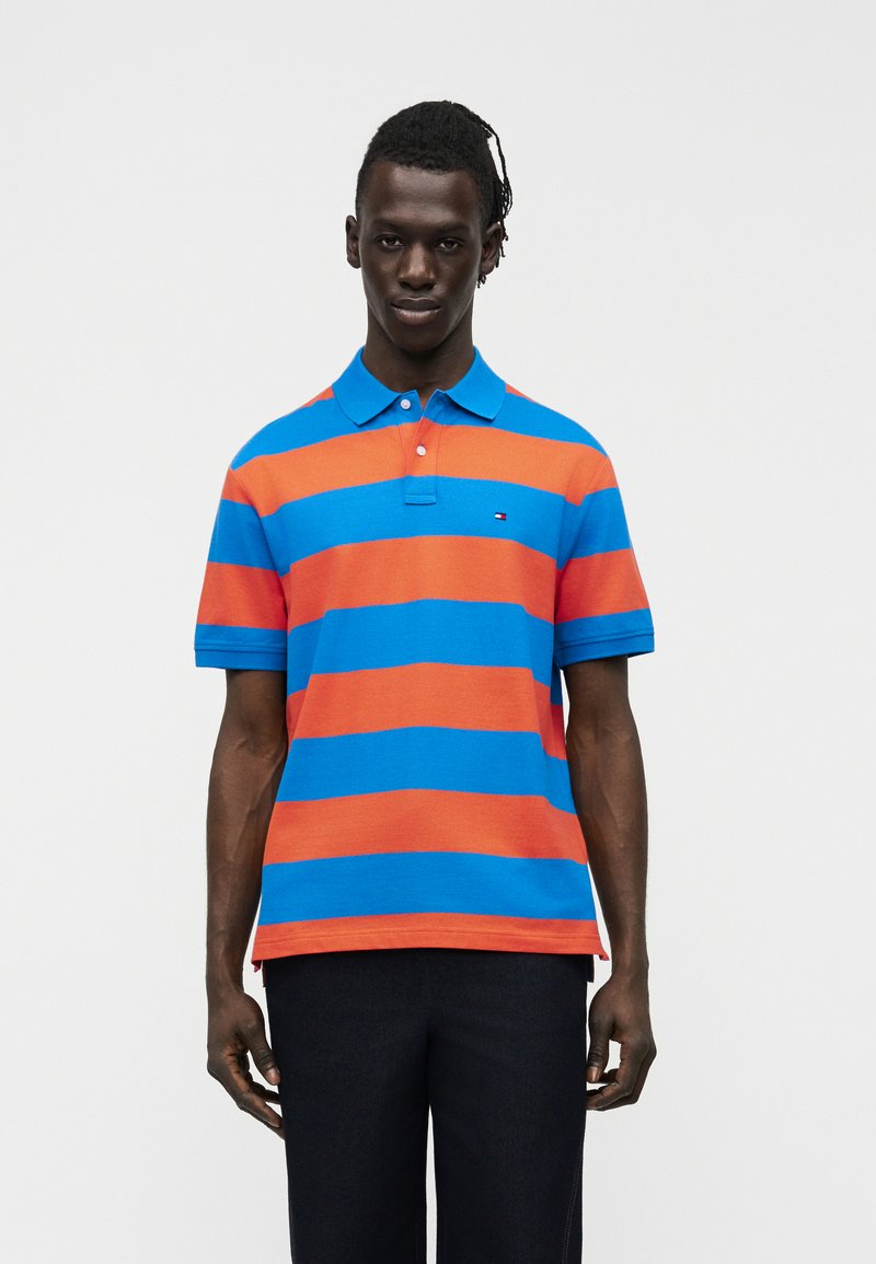 Νεαρός άνδρας με σκούρο δέρμα, φορώντας κοντομάνικο πουλόβερ πόλο με οριζόντιες ρίγες μπλε και πορτοκαλί και μαύρο παντελόνι, στέκεται μπροστά σε απλό φόντο.