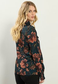 Donkergroene bloes met grote oranje en roze bloemen, gemaakt van zachte stof, met lange mouwen enGathered cuffs, en een kraag. 