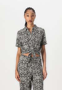 Blouse à manches courtes à motifs noir et blanc avec un nœud à l'avant, ornée de designs géométriques, associée à un pantalon ample assorti.