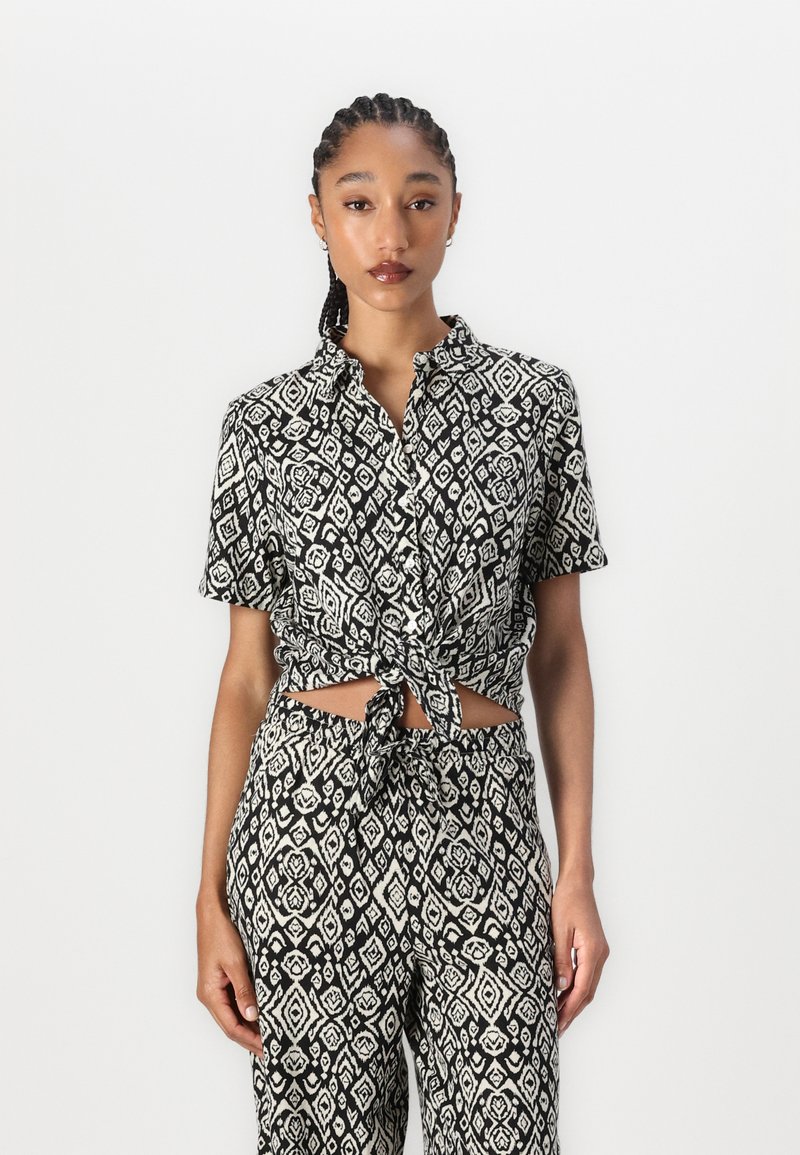 Blouse à manches courtes à motifs noir et blanc avec un nœud à l'avant, ornée de designs géométriques, associée à un pantalon ample assorti.