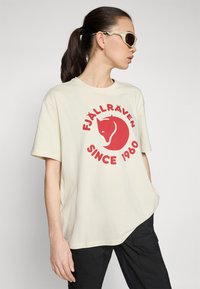 Krém színű pamut póló, piros logóval, amelyen egy róka és a "FJÄLLRÄVEN SINCE 1960" felirat látható. Lazán szabott, rövid ujjú.