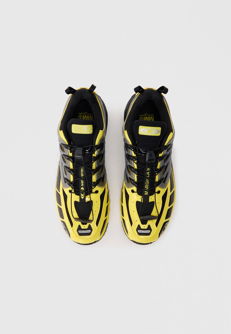 Chaussures de sport noires et jaunes avec une tige en maille, un système de laçage minimal, des semelles en caoutchouc et des découpes de ventilation distinctives sur les côtés.