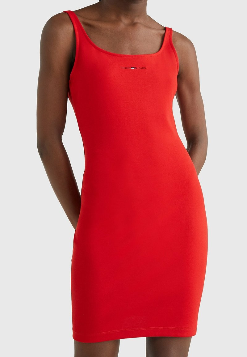 Femme portant une robe rouge moulante sans manches avec un décolleté carré et un petit logo de marque sur la poitrine, sur un fond clair uni.