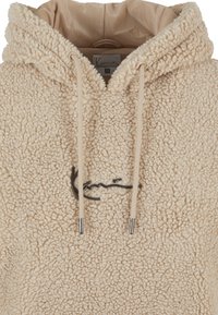 Béžová sherpa fleecová mikina s kapucňou, s kovovými zakončeniami na sťahovacích šnúrkach a malým čiernym podpisovým logom na hrudi.