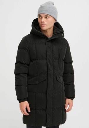 Parka acolchada negra con cremallera frontal, bolsillos con botones a presión, textura acolchada y capucha ajustable; llevada con un gorro de punto gris claro.