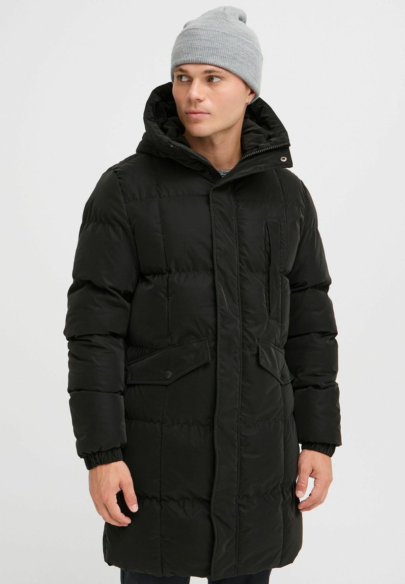Parka rembourrée noire avec une fermeture éclair à l'avant, des poches à bouton-pression, une texture matelassée et une capuche réglable ; portée avec un bonnet en tricot gris clair.