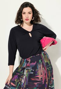 Musta pitkähiha-toppi, jossa on avainaukko, yhdistetty värikkäisiin poimuisiin hameeseen, jossa on abstrakteja kuvioita, ja vaaleanpunaiseen käsilaukkuun.