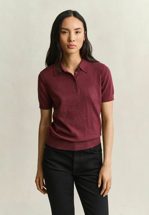 Frau mit langen dunklen Haaren, die ein kurzärmeliges bordeauxfarbenes Poloshirt und schwarze Hosen trägt, steht vor einem schlichten hellen Hintergrund.