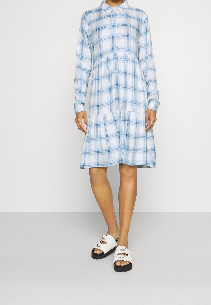 Vestito a camicia a quadri blu e bianchi con colletto, maniche lunghe e orlo a strati. Indossato con sandali bianchi con suole nere e hardware decorativo.