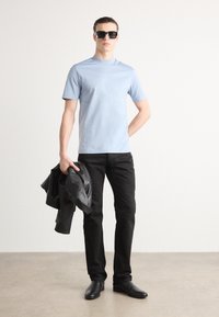 Lys blå kortermet t-skjorte, svarte slim-fit bukser, svart skinnjacket holdt, og mørke solbriller, alt mot en ensfarget bakgrunn.