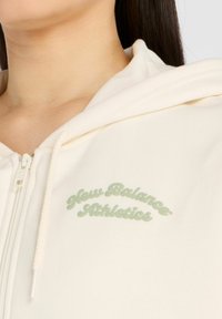 Haut zippé crème en tissu doux, avec un logo New Balance Athletics vert sur la poitrine et une capuche à cordon.