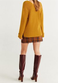 Frau in einem senfgelben Pullover, kariertem Rock in Orange und Braun und knielangen braunen Lederstiefeln, die mit dem Rücken zu uns steht.