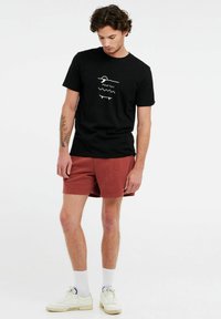 T-shirt nera in cotone con logo grafico bianco e linee curve. Abbinata a pantaloni corti color ruggine e sneakers bianche con suola spessa.