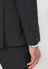 Dunkelgraue Anzugjacke aus strukturiertem Stoff, eng anliegendes Design, vier schwarze Knöpfe am Ärmel und ein sichtbares weißes Hemdärmelloch darunter.