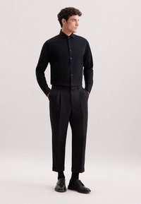 Camicia nera attillata a maniche lunghe con chiusura a bottoni, abbinata a pantaloni neri a pieghe a vita alta e scarpe nere con lacci.