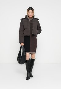 Doudoune brune courte avec deux poches avant, portée sur une robe color-block marron foncé et clair. Bottes noires jusqu'au genou et un sac noir.