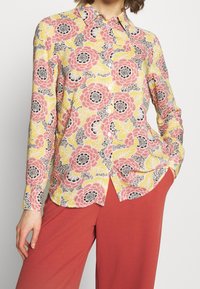 Camisa de botones floral en amarillo con patrones en rosa, negro y blanco. Mangas largas, con cuello, dobladillo curvado. Combinada con pantalones rosa sólidos.