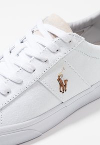 Baskets en toile blanches avec une surface texturée, dotées de lacets blancs, d'une languette en daim beige et d'un logo brodé marron d'un joueur de polo.