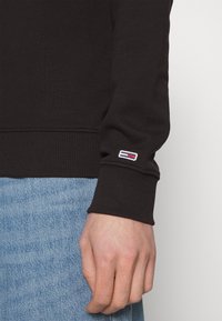 Felpa nera con polsini e vita a costine. Presenta una piccola toppa con logo con accenti rossi, bianchi e blu sulla manica. Jeans in denim visibili.