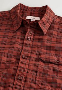 Punane ja must ruuduline nööbitav särk, mis on valmistatud puuvillast, struktuursed krae, rinnatasku ja pruunid nööbid.