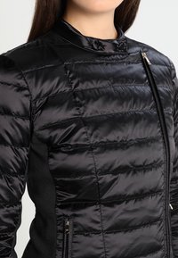 Femme portant une veste matelassée noire avec un col montant et une fermeture éclair diagonale, gros plan sur le haut du torse et le cou.