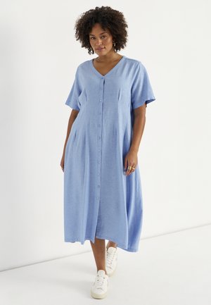 Kobieta o kręconych włosach, ubrana w jasnoniebieską sukienkę midi zapinaną na guziki z przodu i białe trampki, stojąca na białym tle.