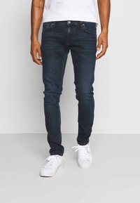 Mörkblå denimjeans med slim fit, som har fem fickor, dragkedja och diskret blekning. Stylingen kompletteras med vita sneakers.