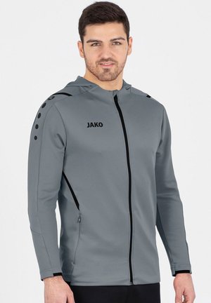 Uomo con felpa grigia con zip JAKO con dettagli neri, in piedi davanti a uno sfondo chiaro e uniforme, che guarda avanti e sorride leggermente.