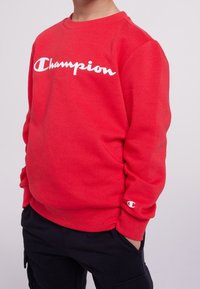 Bambino che indossa un luminoso maglione Champion rosso con il logo bianco sul petto e un piccolo logo sulla manica, con le mani nelle tasche dei pantaloni neri.