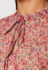 Blommig blus med rynkad halsringning, nyckelhålsdetalj och band. Materialet visar en blandning av röda, rosa och blå blommönster på en neutral bakgrund.