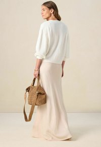 Vrouw in een witte gestructureerde trui en een beige lange rok, met een bruine geperforeerde handtas met een schouderband, zijwaarts gekeerd.