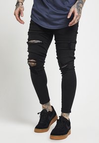 Jeans ajustados negros rasgados con múltiples parches desgastados y un corte slim. Combinados con zapatillas negras con una suela texturizada.