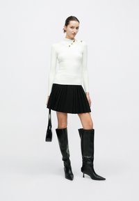 Λευκό ριμπ μακρυμάνικο τοπ με ψηλό γιακά και διακοσμητικά κουμπιά, συνδυασμένο με μαύρη πλισέ μίνι φούστα και μαύρες μπότες μέχρι το γόνατο.