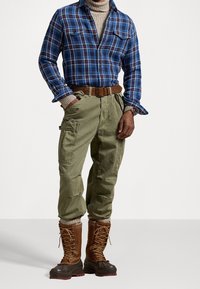 Camicia di flanella a quadri blu e rossi, pantaloni cargo oliva, cintura in pelle marrone e stivali impermeabili marroni con design con lacci.