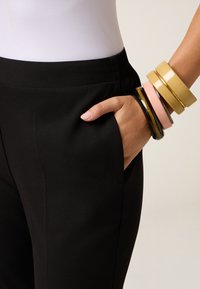 Pantaloni neri con una tasca laterale, accompagnati da diversi braccialetti colorati in giallo, rosa e nero su un polso. Tessuto di texture liscia.