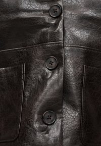 Κοντινό πλάνο σκούρου καφέ δερμάτινου μπουφάν με μεγάλα μαύρα κουμπιά και δύο ραμμένες μπροστινές τσέπες.