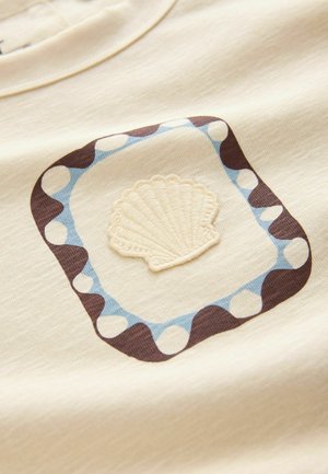 Beige stof med et indrammet design, der har en cremefarvet broderet muslingeskal i midten og en blå og brun prikket kant.