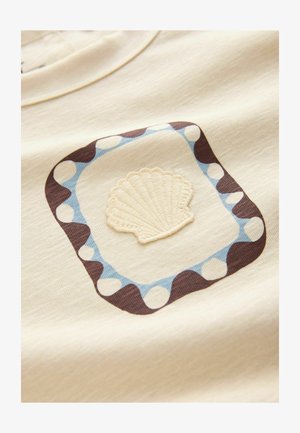 Tela beige con un diseño enmarcado que presenta una concha marina bordada de color crema en el centro y un borde punteado azul y marrón.