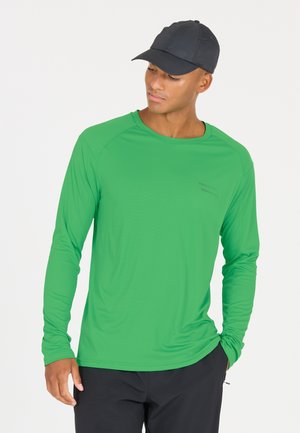 Leichtes Langarmhemd in leuchtendem Grün, mit strukturiertem Stoff und Rundhalsausschnitt. Schwarze Mütze, schwarze Hose. Minimalistische Akzente auf dem Hemd.