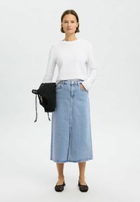 Falda larga de mezclilla azul claro con costura frontal y un ligero desgaste, combinada con una blusa blanca de manga larga y un bolso negro de tela tejida. Zapatos planos negros.