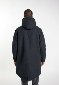 Fekete kapucnis parka, hosszított hátrésszel, rugalmas mandzsettákkal és húzózsinóros alsó résszel. Simaság jellemzi, minimális dizájn, nem látható mintázattal.
