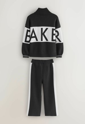 Crno-bijela pletena majica s turtleneckom i širokim rukavima, s natpisom "BAKER". Uparena s crnim hlačama koje imaju bijele bočne pruge.