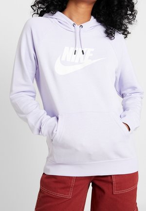 Kobieta nosząca jasnofioletową bluzę z kapturem Nike z przednią kieszenią i troczkami, w połączeniu z czerwonymi spodniami z widocznymi szwami.